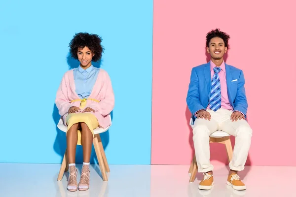 Smiling african american couple sitting on chairs against pink and blue wall backdrop — Stock Photo