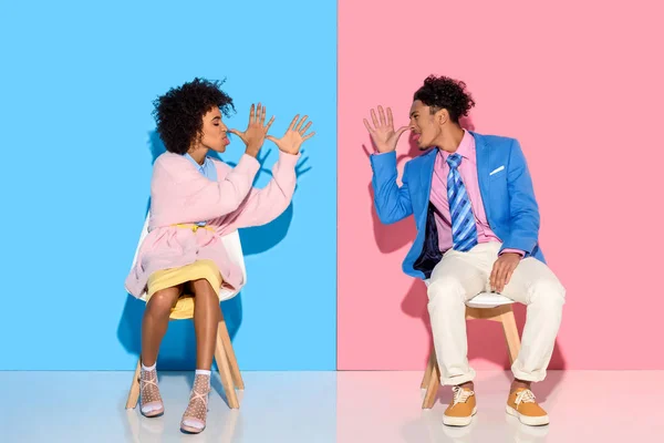 Couple afro-américain grimace les uns aux autres tout en étant assis sur des chaises contre fond de mur rose et bleu — Photo de stock