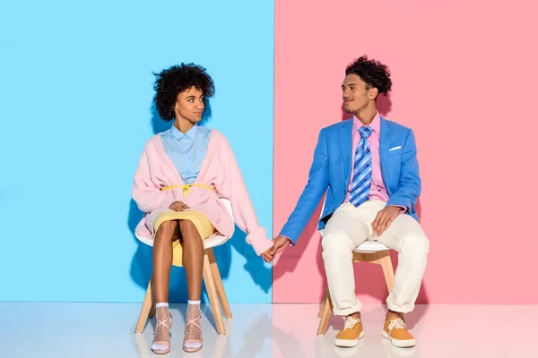 African american couple holding hands while sitting on chairs against pink and blue wall backdrop — Stock Photo