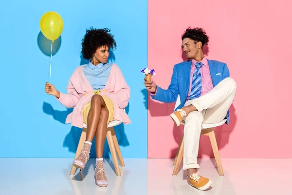 African american man presenting flowers in ice cream cone to girlfriend while sitting on chairs against pink and blue wall backdrop — Stock Photo