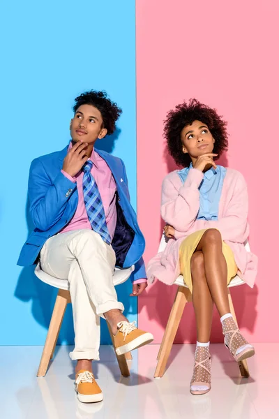 Africano americano pensativo pareja sentado en sillas contra rosa y azul pared telón de fondo - foto de stock