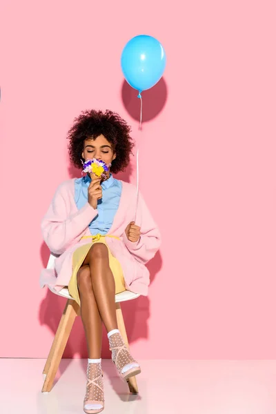 Mujer afroamericana con flores en cono de helado y globo en las manos sobre fondo de pared rosa - foto de stock