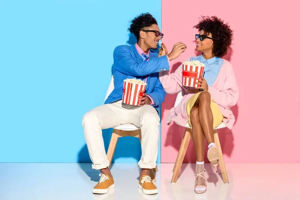 Joven africano americano pareja sentado en sillas y comer palomitas de maíz contra rosa y azul pared - foto de stock