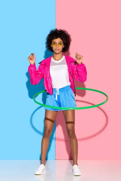 Deportiva mujer posando mientras hace ejercicio con aro y mirando a la cámara sobre fondo rosa y azul - foto de stock