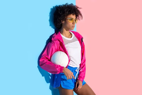 Junges Mädchen mit Brille, Ball in der Hand und wegschauend auf rosa und blauem Hintergrund — Stockfoto