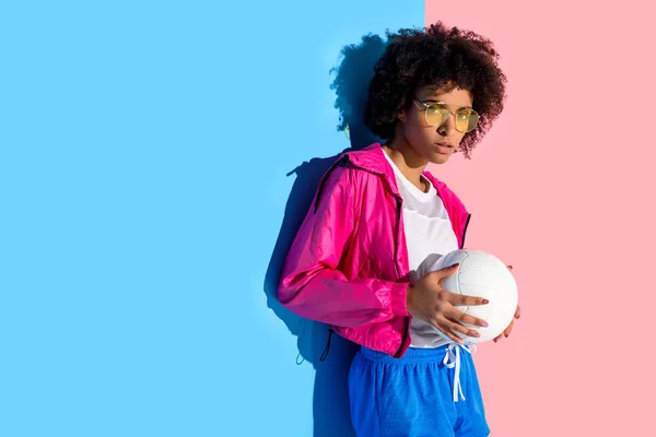 Молода дівчина в окулярах тримає м'яч і дивиться на камеру на рожевому і синьому фоні — стокове фото