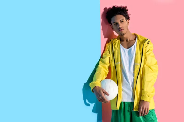 Joven guapo sosteniendo la pelota en la mano y mirando hacia otro lado sobre fondo rosa y azul - foto de stock