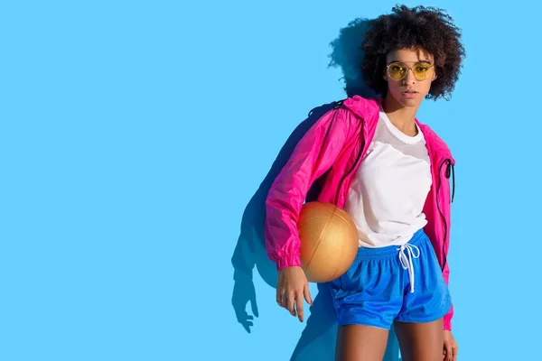 Молодая девушка в очках держит баскетбольный мяч и смотрит в сторону на синем фоне — стоковое фото