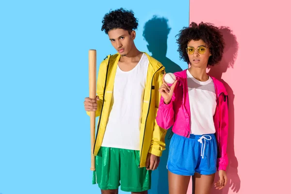 Jeune couple attrayant avec équipement de baseball dans les mains regardant la caméra sur fond rose et bleu — Photo de stock