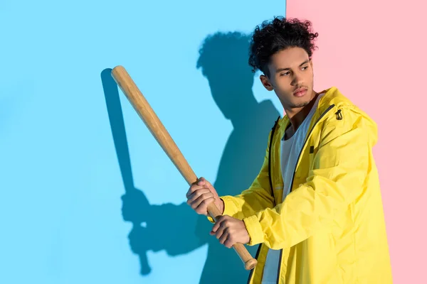 Bello giovane uomo che tiene in mano la mazza da baseball in procinto di calciare e distogliere lo sguardo su sfondo rosa e blu — Foto stock