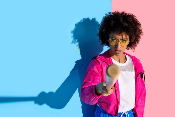 Jeune fille afro-américaine en colère pointant à la caméra avec batte de baseball sur fond rose et bleu — Photo de stock