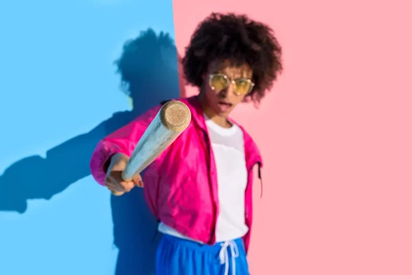 Jovem menina americana africana irritada apontando na câmera com bastão de beisebol no fundo rosa e azul — Fotografia de Stock