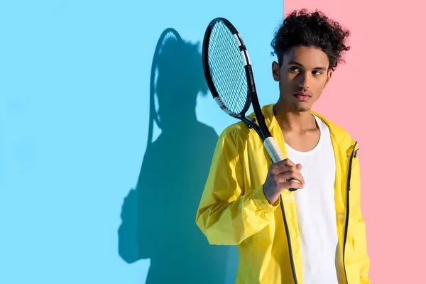 Joven chico afroamericano brillante sosteniendo raqueta de tenis y mirando hacia otro lado en el fondo rosa y azul - foto de stock