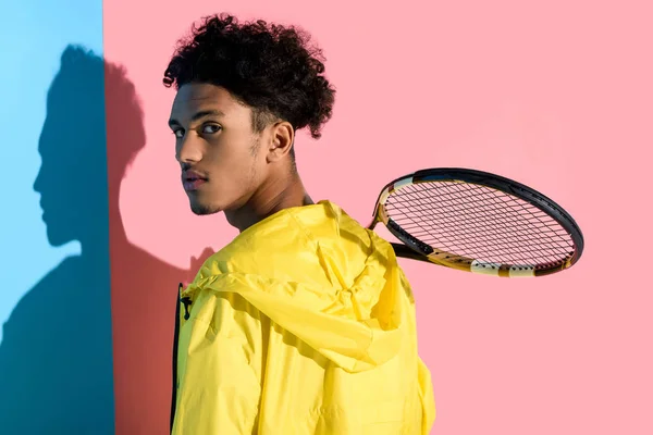 Молодий яскравий афроамериканський хлопець тримає тенісну ракетку на плечі на рожевому і синьому фоні — стокове фото