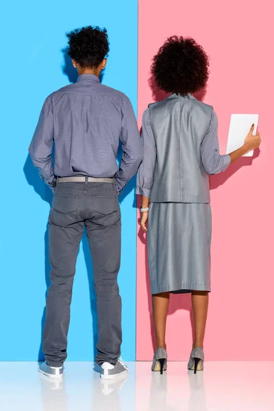 Vista posteriore di giovane uomo d'affari africano amerciano in piedi da donna d'affari con tablet in mano su sfondo rosa e blu — Foto stock