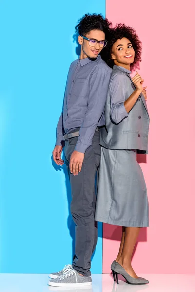 Young african amercian smiling couple in grey suits standing back to back on pink and blue background — Stock Photo