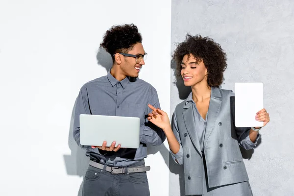 Jovem empresário afro-americano com laptop e empresária com tablet na mão sobre fundo cinza e branco — Fotografia de Stock