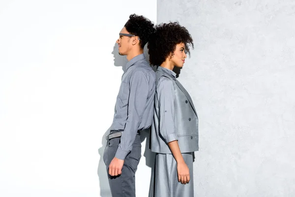 Молодой афроамериканец улыбающаяся пара в серых костюмах, стоящая спиной к спине на сером и белом фоне — стоковое фото