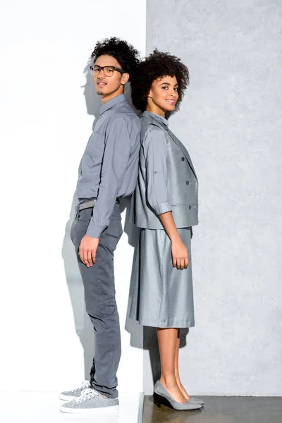 Young african amercian smiling couple in grey suits standing back to back and looking at camera on grey and white background — Stock Photo