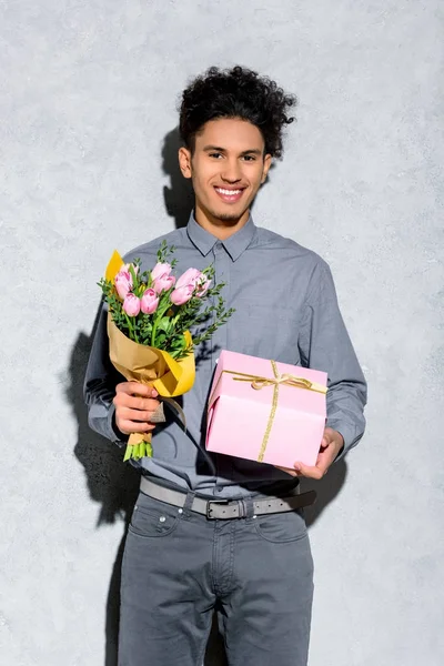 Junger afrikanisch-amerikanischer Mann mit Blumenstrauß und Geschenk auf grauem Hintergrund — Stockfoto