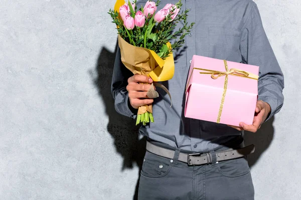 Крупный план человека, держащего букет цветов и подарок на сером фоне — стоковое фото