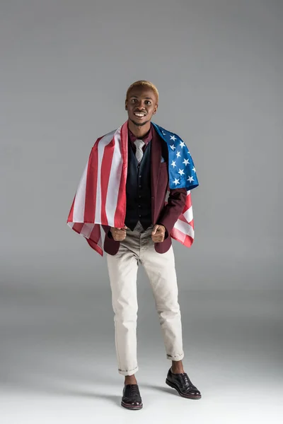 Усміхнений афроамериканський чоловік з американським прапором на плечах дивиться на камеру ізольовано на сірому — стокове фото
