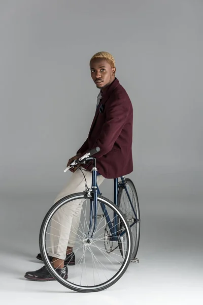 Ernster afrikanischer Mann sitzt auf dem Fahrrad und blickt in die Kamera auf grauem Hintergrund — Stockfoto