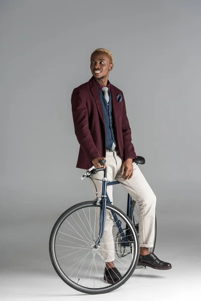 Улыбающийся африканский мужчина сидит на велосипеде и смотрит в сторону на сером фоне — стоковое фото