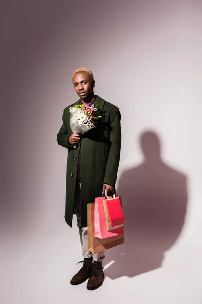 Elegante joven afroamericano hombre sosteniendo ramo de flores y brillantes bolsas de compras - foto de stock