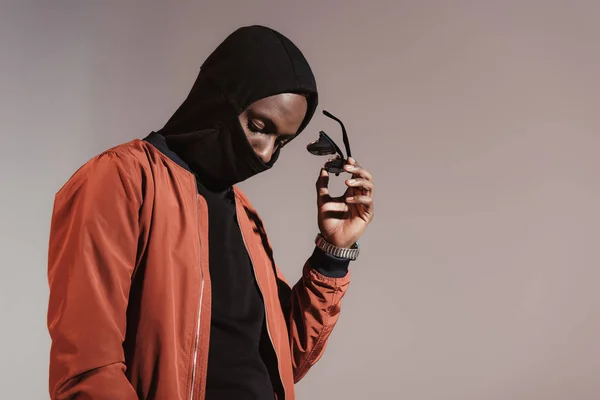 Elegante jovem afro-americano vestindo capuz com máscara facial e tomando de seus óculos de sol isolados no fundo de luz — Fotografia de Stock