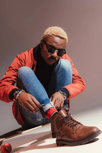 Stylish young african american skater sits on longboard and straightens his jeans — Stock Photo