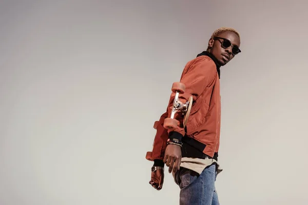 Stylish young african american skateboarder holding longboard behind his back isolated on light background — Stock Photo