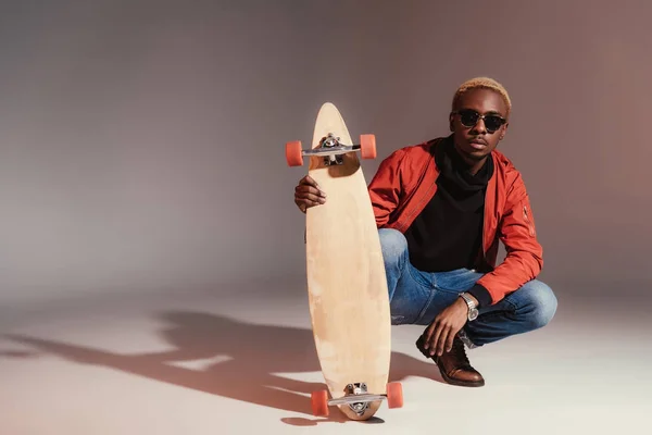 Elegante joven afroamericano skateboarder sentado y sosteniendo longboard - foto de stock