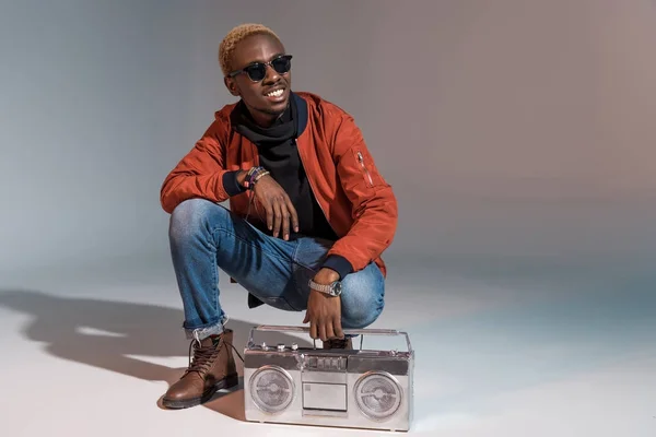 Fröhlich stylische junge afrikanisch-amerikanische Mann hockt mit silbernem Tonbandgerät auf grau — Stockfoto