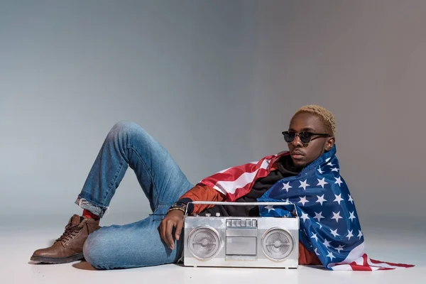 Jeune homme afro-américain avec nous drapeau sur les épaules couché avec magnétophone argent sur gris — Photo de stock