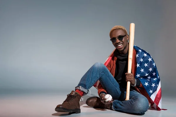 Fröhlicher junger afrikanisch-amerikanischer Mann mit unserer Fahne auf den Schultern, Baseballschläger und Ball auf grau — Stockfoto