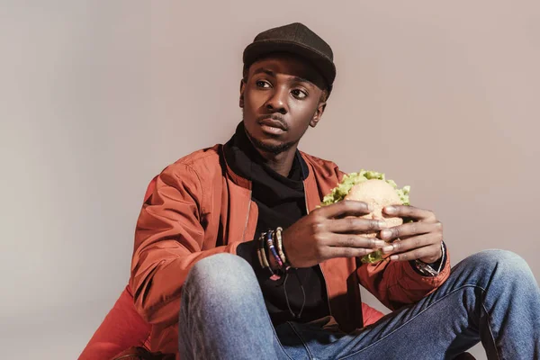 Joven afroamericano hombre comiendo hamburguesa y mirando hacia otro lado aislado en gris — Stock Photo
