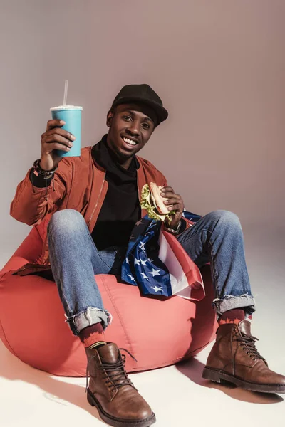 Happy young african american man eating hamburger and drinking from paper cup while sitting on bean bag chair in studio — Stock Photo