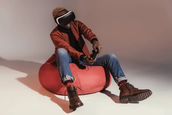 Young african american man in virtual reality headset playing with joystick while sitting on bean bag chair in studio — Stock Photo