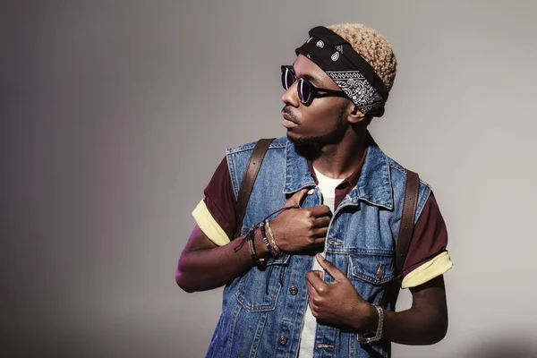 Porträt eines eleganten jungen afrikanisch-amerikanischen Mannes mit Sonnenbrille und Lederrucksack, der grau wegschaut — Stockfoto