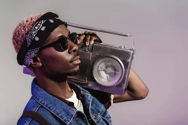 Стильний молодий афроамериканський чоловік в сонцезахисних окулярах тримає магнітофон на плечі і дивиться ізольовано на сірий — стокове фото