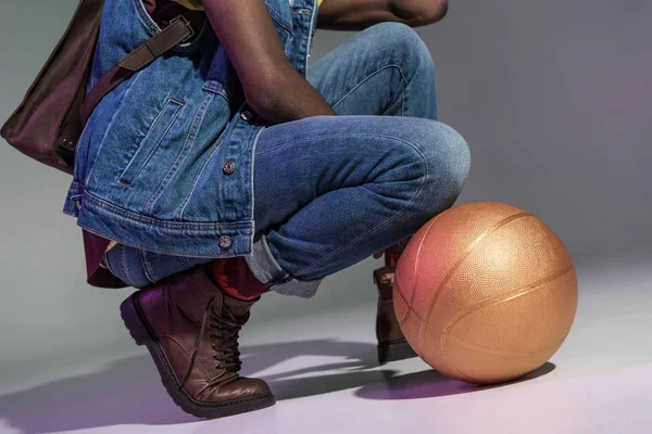 Colpo ritagliato di uomo afroamericano accovacciato con pallone da basket dorato su grigio — Foto stock