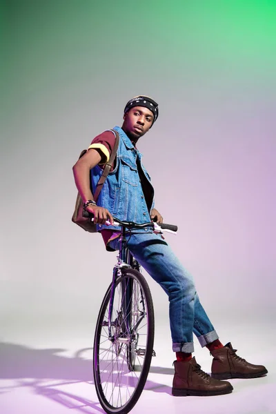 Hombre afroamericano joven y elegante sentado en bicicleta y mirando hacia otro lado en gris - foto de stock