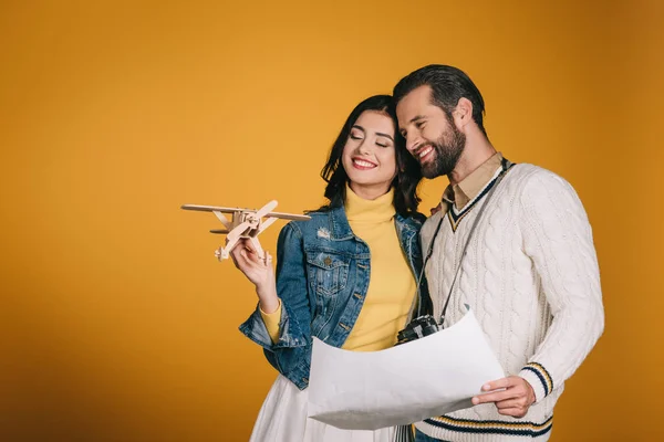 Sonriente pareja mirando avión de madera aislado en amarillo - foto de stock