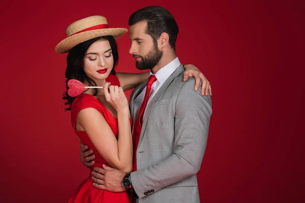 Freundin hält herzförmigen Lutscher und umarmt Freund isoliert auf rot — Stockfoto