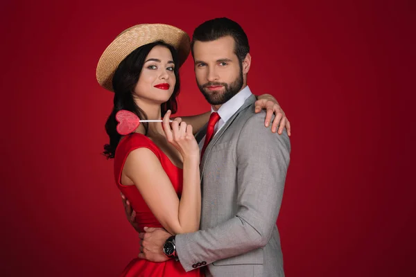 Girlfriend holding heart shaped lollipop and hugging boyfriend isolated on red — Stock Photo