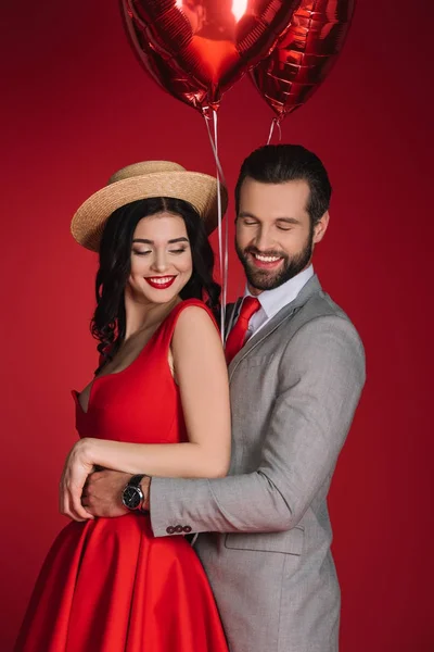 Sonriente elegante pareja con globos rojos aislados en rojo - foto de stock