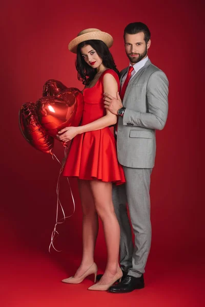 Attractive couple with heart shaped balloons looking at camera on red — Stock Photo