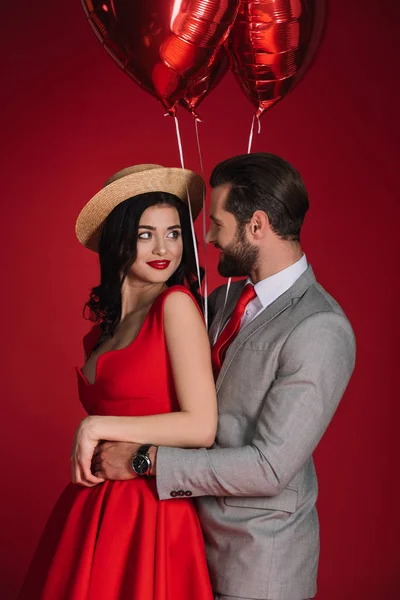 Stylisches Paar mit roten Luftballons, die sich isoliert auf Rot umarmen — Stockfoto