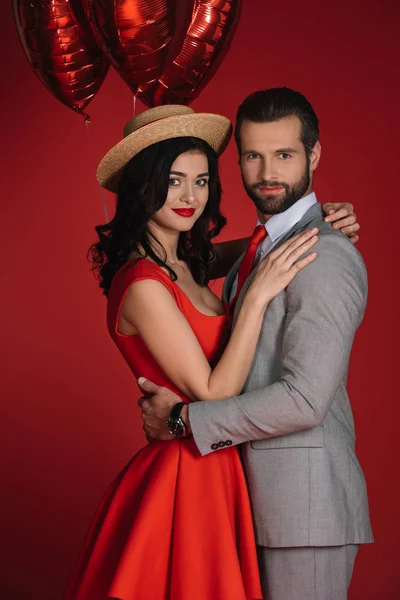 Stilvolles Paar mit roten Luftballons, das isoliert auf rot in die Kamera blickt — Stockfoto